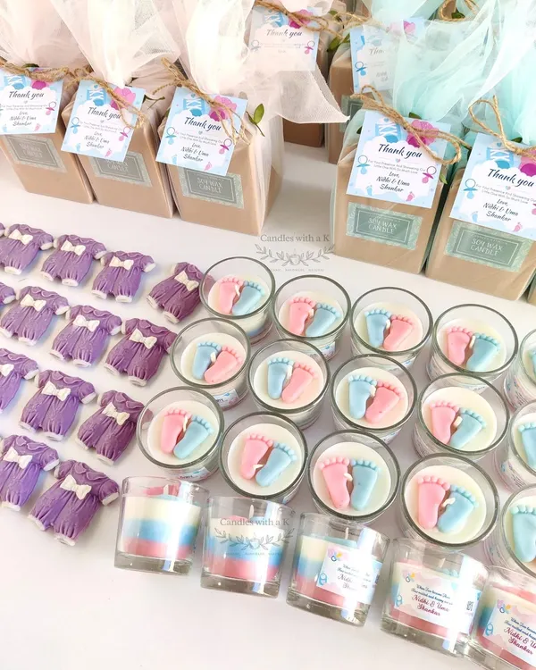 A complete baby shower return gift set, featuring a layered votive candle with tiny pink and blue baby feet, and a matching baby onesie wax sachet.