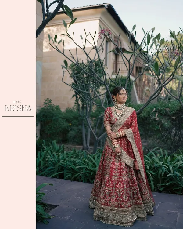 A vision of bridal elegance. This full-length portrait captures the bride in her magnificent red Sabyasachi lehenga, standing gracefully amidst lush greenery.