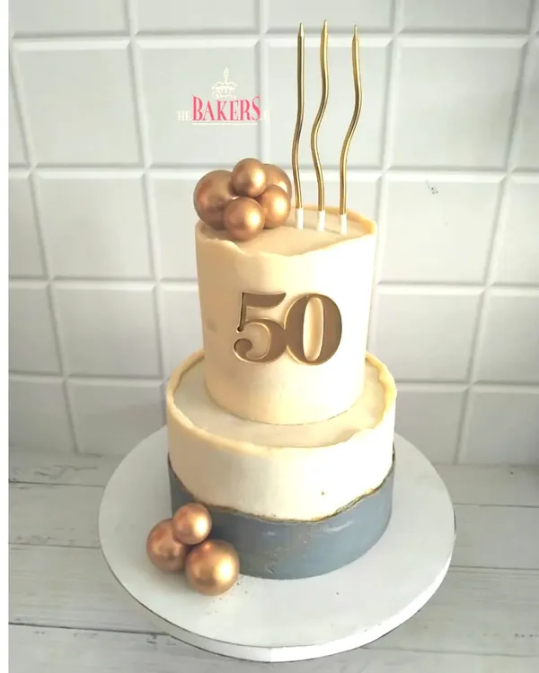 An elegant two-tier cake for a 50th birthday celebration. This no-fondant design features a modern, minimalist aesthetic with a concrete-grey bottom tier and a clean white top tier, accented with gold spheres and a bold "50" topper.