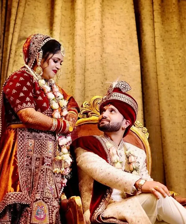 A regal wedding portrait of the couple. The groom is seated while the bride stands beside him, creating a classic and royal composition.