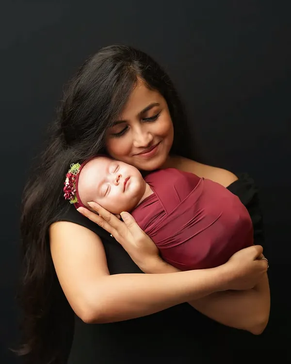 A mother with long dark hair, dressed in black, holds her sleeping newborn close to her chest. The baby is swaddled in a deep red wrap, creating a beautiful color contrast against the dark backdrop.