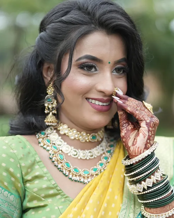 A happy bride adjusts her nath, giving a beautiful view of her layered Kundan necklace set with sea-green stones and matching jhumka earrings.