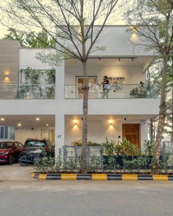 The facade of Villa 17 in Bangalore. This two-story home features a clean white exterior, a glass balcony railing, and strategically placed lighting, softened by a ledge of green creepers for a touch of nature.