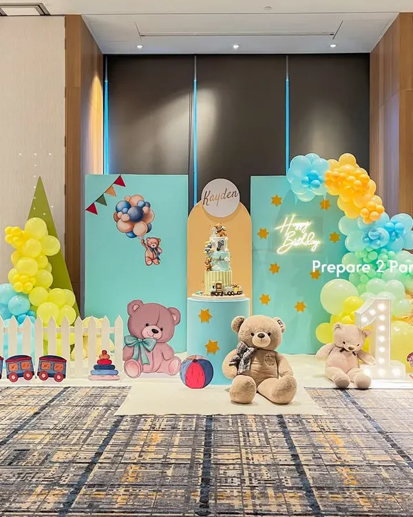 A cheerful and playful first birthday party setup in a large hall. The theme features pastel blue and yellow backdrops, a white picket fence, cute teddy bear cutouts, and a light-up marquee number "1".