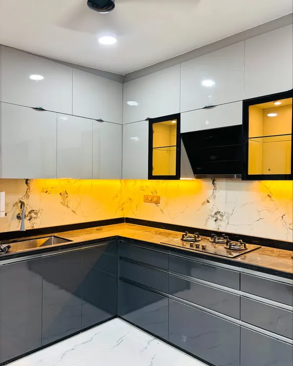 A modern L-shaped modular kitchen featuring a two-tone combination of high-gloss grey and white cabinets. This design includes handleless drawers, a built-in chimney, and warm under-cabinet LED lighting against a marble-patterned backsplash.