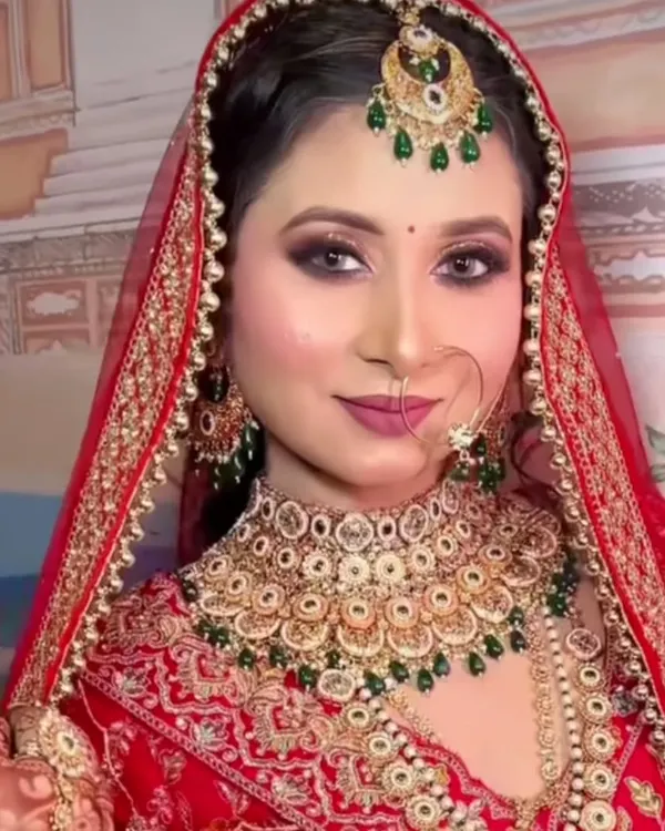 A full view of a classic red bridal look available for rent. This set includes a gold-plated choker with green accents, a matching maang tikka, and a simple nath.
