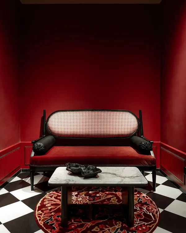 A statement piece from our House of Goya collection. This classic daybed features a black-lacquered frame, a cane back, and rich red velvet upholstery. It's set in a dramatic, color-drenched room with a checkerboard floor.