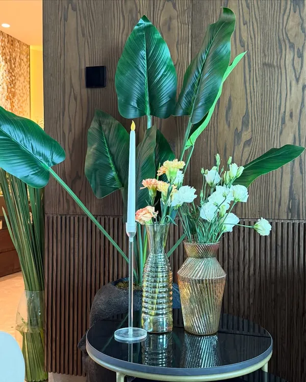 For this housewarming in a luxurious Noida home, we placed a simple yet elegant arrangement on a side table. The combination of fresh white flowers, large green leaves, and a single candle complements the modern wood-paneled wall perfectly.