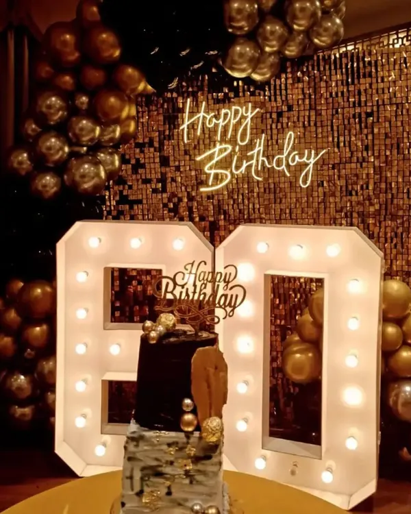 This 60th birthday photo corner was designed for maximum impact. It features a gold sequin shimmer wall, a 'Happy Birthday' neon sign, and oversized light-up '60' numbers, complemented by a classic black and gold balloon garland.