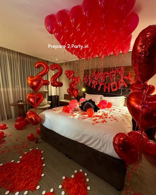 A classic romantic birthday room decoration in a hotel suite. The setup features red heart-shaped foil balloons, number balloons indicating the age, "Happy Birthday" lettering above the bed, and a floor decorated with rose petal hearts.