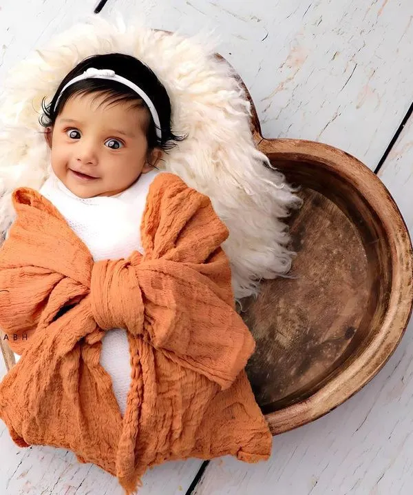 A precious moment of connection as this wide-eyed baby smiles from a cozy, heart-shaped wooden bowl. This is the kind of spontaneous magic we love to capture during our at-home newborn photoshoots.