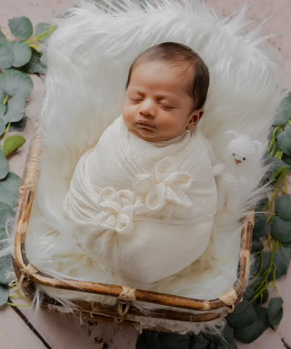 A perfect little bundle of joy. This newborn is peacefully asleep in a rustic basket, swaddled in white on a fluffy fur blanket and surrounded by eucalyptus leaves.
