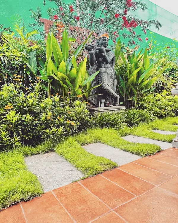 A statue of Krishna playing the flute is nestled among lush tropical foliage and vibrant red flowers. The pathway, made of stone pavers interspersed with grass, leads to this spiritual alcove, creating a sacred pause within the garden.