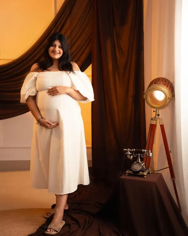 A classic studio portrait with a vintage touch. The mom-to-be looks radiant in a white off-the-shoulder dress, complemented by the warm lighting and antique props.