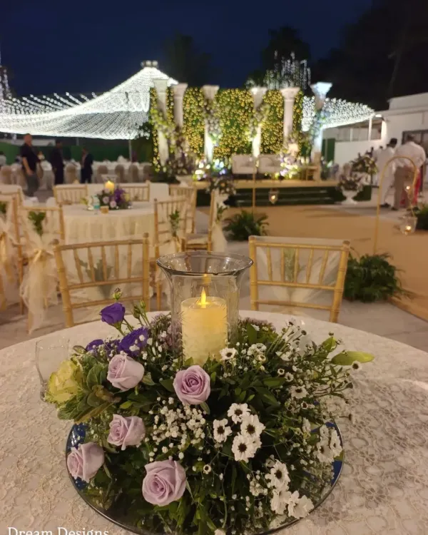 A close-up of a classic and elegant centerpiece. A pillar candle in a glass hurricane lamp is surrounded by a delicate arrangement of lavender roses, baby's breath, and daisies, all set on a mirrored base.