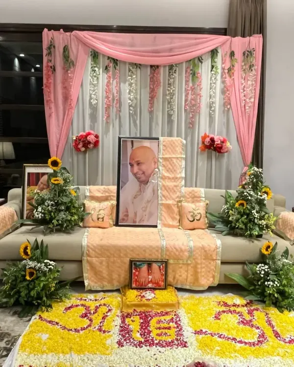 This is a complete view of a Guruji Satsang I organized. The main seating area features a portrait of Guruji, decorated with soft pink drapes and floral arrangements. In front, a large, intricate floral rangoli with the 'Om' symbol creates a powerful focal point for meditation and prayer.