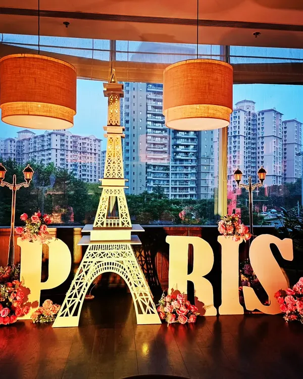 A stunning Paris-themed setup against a city skyline. The decor features a large Eiffel Tower cutout, 'PARIS' letters, and beautiful floral arrangements, perfect for a chic and sophisticated event.