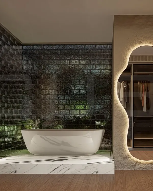 A luxurious bathroom featuring a freestanding bathtub set on a raised marble platform against a textured glass brick wall. The organically shaped, backlit mirror adds a soft, ambient glow and serves as a unique design element.