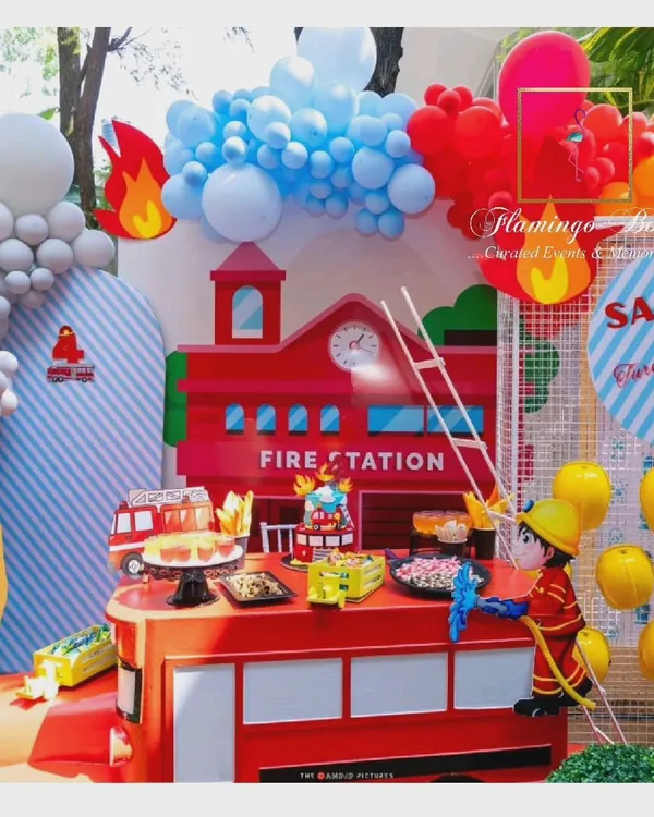 The main setup for a firefighter-themed birthday, featuring a custom fire station backdrop and a dessert table designed to look like a fire truck. Balloon arrangements in red, blue, and orange mimic fire and water.