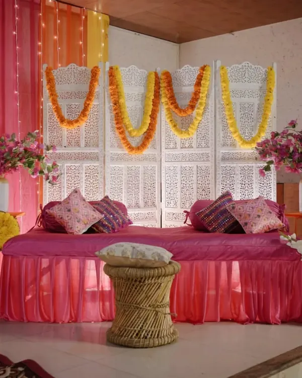 A classic Mehendi seating arrangement featuring a comfortable divan in bright pink, set against a traditional white carved screen and decorated with marigold garlands.
