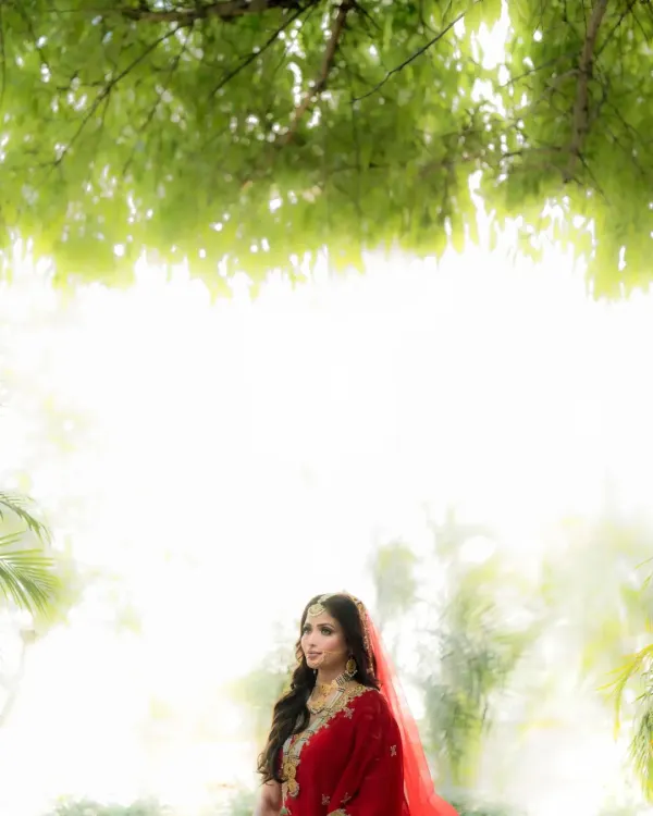 A bride framed by nature, her red lehenga glowing against the soft, diffused light. This is a portrait of pure, ethereal beauty.