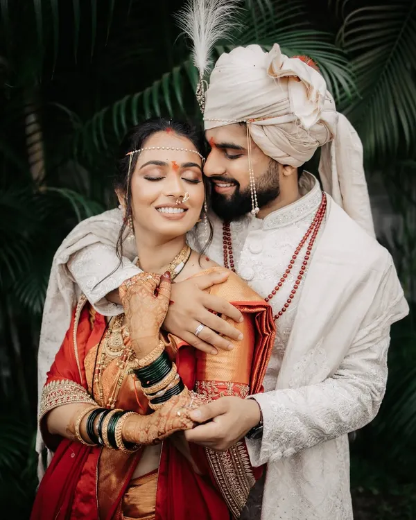 An intimate portrait of a Maharashtrian couple, the groom embracing his bride, who is adorned in a traditional red saree and jewelry.