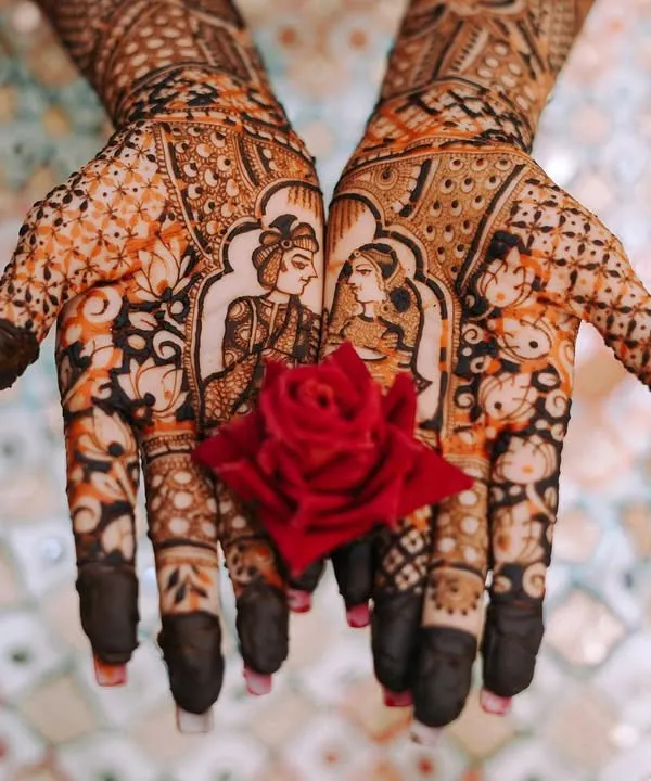 A close-up of the bride's hands showcasing the incredible detail of her mehndi, featuring portraits of the couple. A small red rose adds a beautiful pop of color.