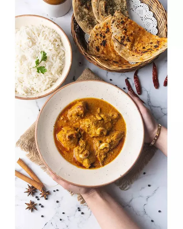 A light and airy composition of Chicken Korma, served with rice and roti. The hands holding the bowl add a human element, making the meal feel warm and inviting.