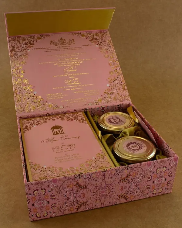 An exquisite pink and gold boxed invitation, perfect for a grand wedding or engagement. The box opens to reveal the invitation details printed on the inside of the lid, with compartments for the card inserts and two jars for sweets or dry fruits.