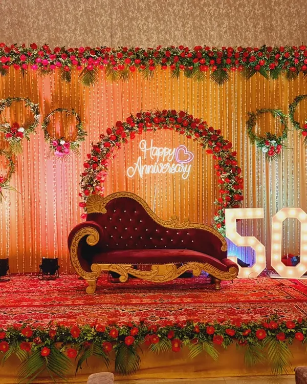 A complete stage setup for a 50th anniversary celebration. This includes a red floral backdrop, a 'Happy Anniversary' neon sign, large '50' marquee numbers, and a classic chaise lounge for seating.