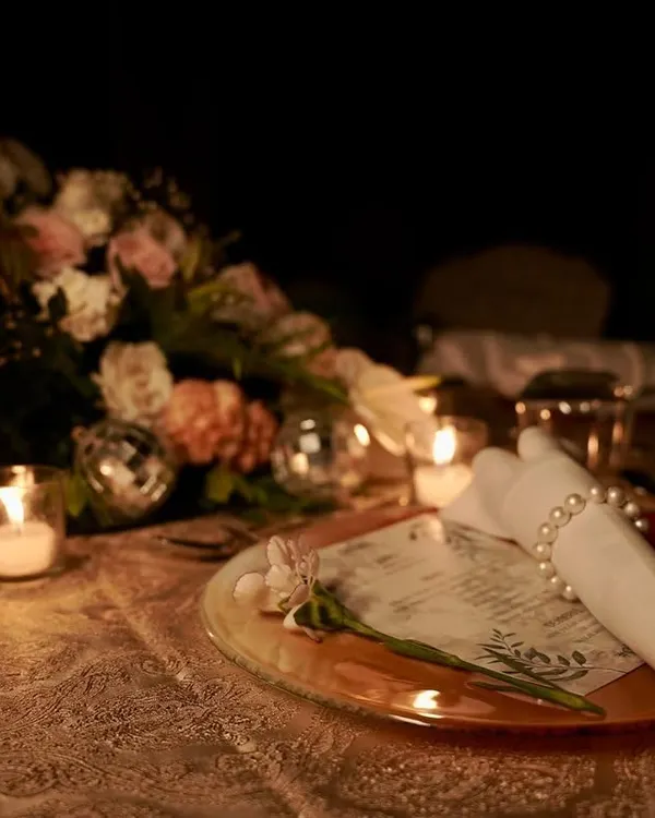 A beautifully set table for an intimate dinner. The place setting features a personalized menu, a pearl napkin ring, and a single flower, all glowing in the warm light of a nearby candle.