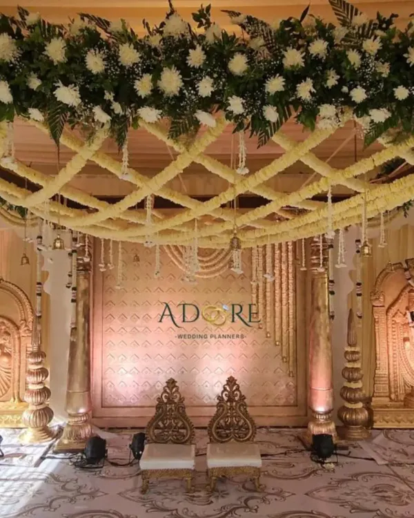 A magnificent golden-themed mandap for a traditional Muhurtham ceremony. The design features a lattice of white flowers on the ceiling, ornate pillars, and idols of deities, creating a sacred and regal space.