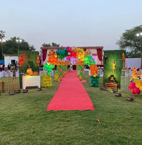 A grand entrance for a jungle party at a farmhouse, featuring a red carpet walkway lined with green hedge walls, animal props, and vibrant balloon arches.