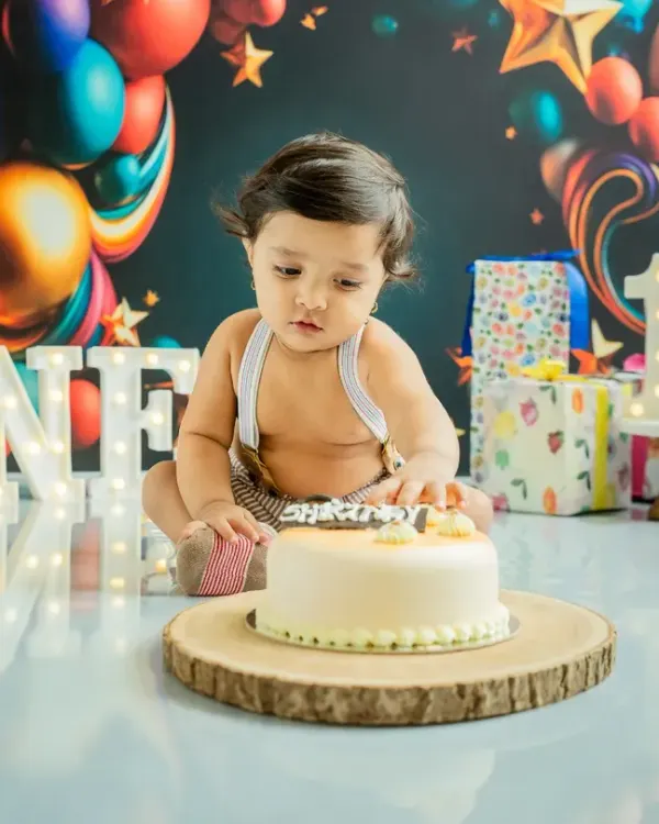 The moment of anticipation just before the smash. This little one is eyeing his first birthday cake in a festive setup with 'ONE' marquee lights and colorful balloons.