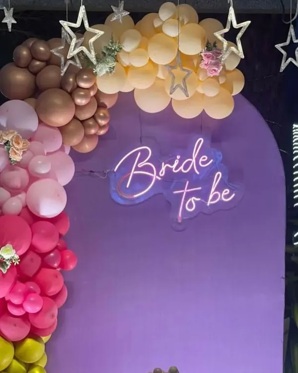 A 'Bride to be' party backdrop with a purple panel, a neon sign, and a colorful balloon garland.