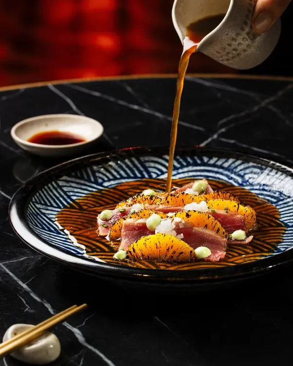 A moody and elegant shot for Akina Mumbai, featuring a tuna dish with a sauce pour. The dark marble tabletop and dramatic lighting highlight the vibrant colors of the food and the precise plating.