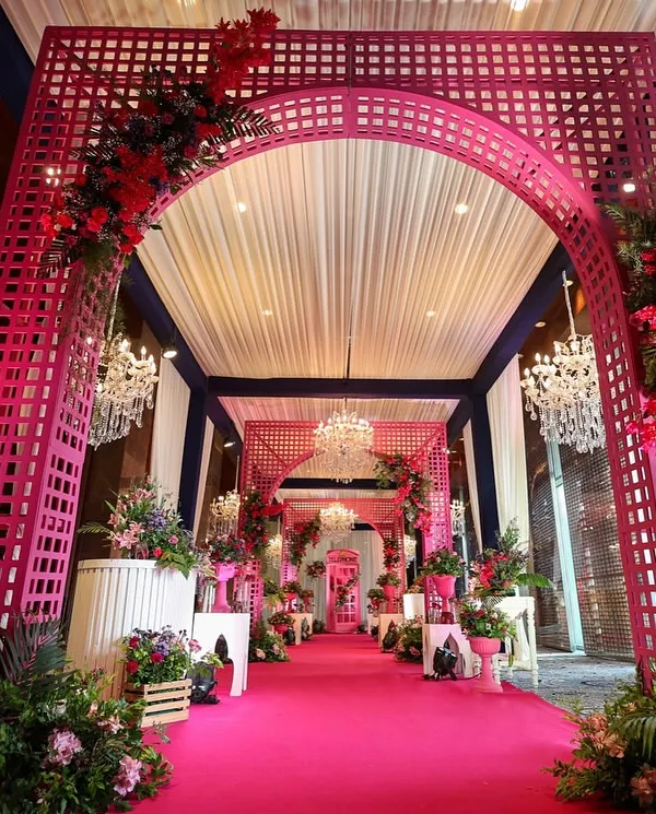 A grand entrance for a Haldi ceremony, designed with a stunning pink lattice archway and a carpeted walkway. The entire space is bathed in shades of pink and red florals, complemented by crystal chandeliers for a touch of luxe.