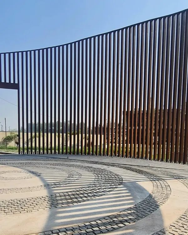 The entrance to a residential development in Raipur, featuring a gracefully curving Corten steel gate. The shadows cast by the vertical slats create a dynamic pattern on the cobblestone driveway.