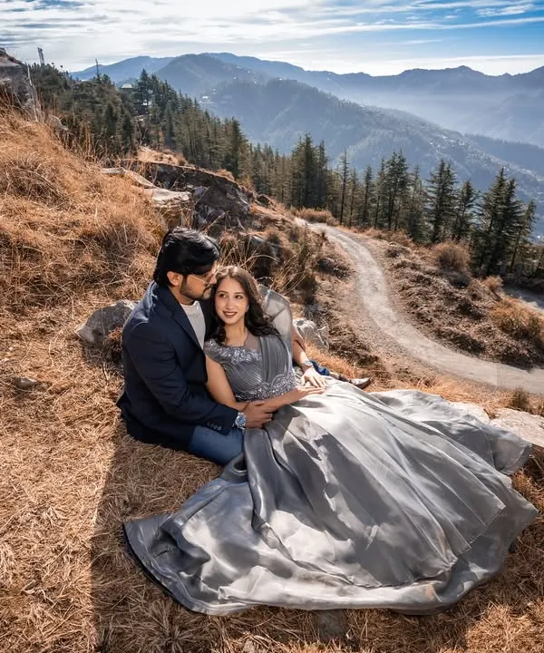 A stunning wide-angle shot of the couple nestled on a mountainside, with layers of Himalayan ranges stretching into the distance. The flowing grey gown adds to the epic feel.