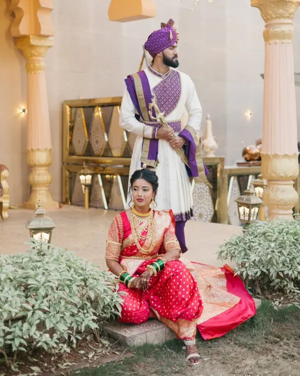 A regal portrait set against a palatial backdrop in Thane. The bride is seated in her vibrant red Nauvari saree, while the groom stands tall in his traditional kurta and pheta, creating a scene of timeless grace.