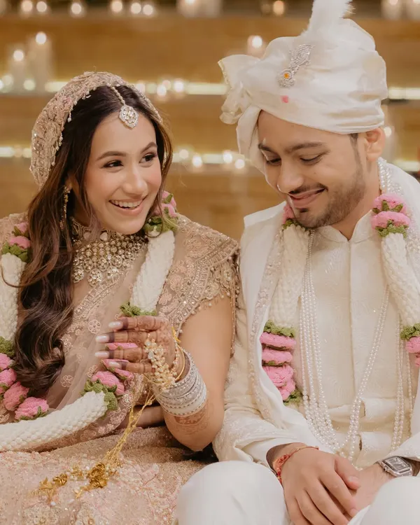 A candid, happy moment from a wedding ceremony. The bride's pastel pink lehenga is paired with a soft, doll-like makeup look, featuring rosy cheeks and a gentle glow that reflects her joy.