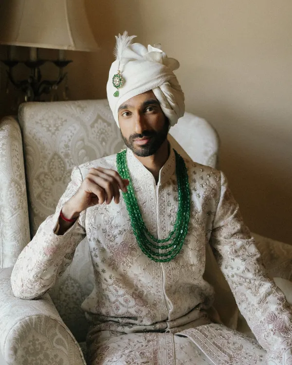 A classic portrait of the groom, Sid, seated in a regal armchair. His emerald green necklace provides a stunning pop of color against his ivory sherwani.
