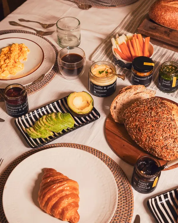 A beautiful breakfast spread featuring scrambled organic eggs, a fresh croissant, sliced avocado, and our artisanal pantry items like hummus, pesto, and jam. This is how we bring our table to your home.