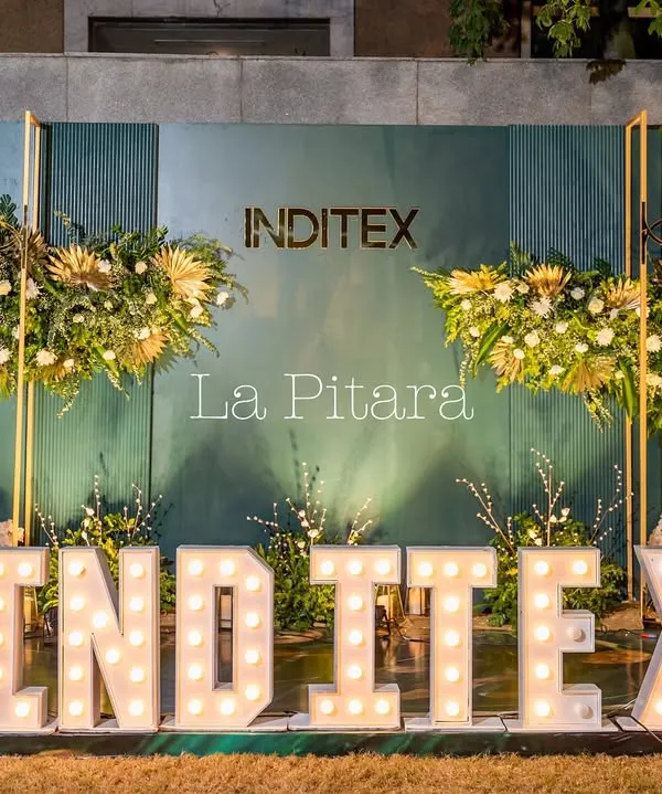 A stunning photo backdrop designed for the Inditex Diwali party, featuring large marquee letters of the company name. The setup is framed by modern gold structures with lush floral arrangements, creating a perfect spot for corporate branding and guest photos.