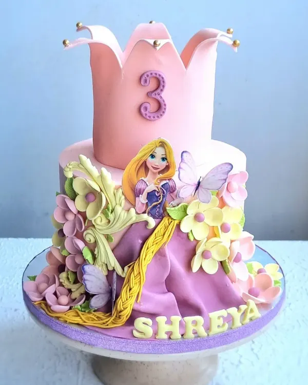 A magical Rapunzel-themed cake for a third birthday. This design features a crown-shaped top tier, a fondant Rapunzel figure, her long braided hair, and beautiful sugar flowers and butterflies.