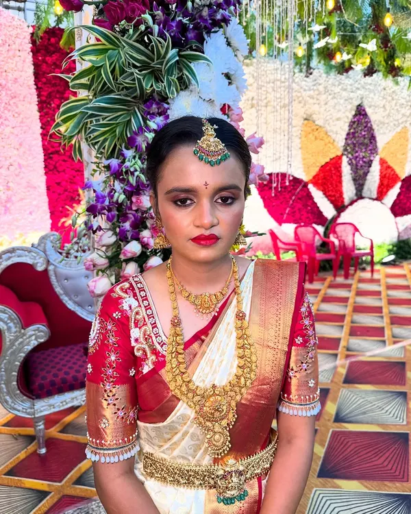 A classic muhurtham look for Priyanka in her beautiful white and red Kanjeevaram saree. The makeup features a bold red lip and defined eyes, perfectly complementing her traditional gold jewellery.