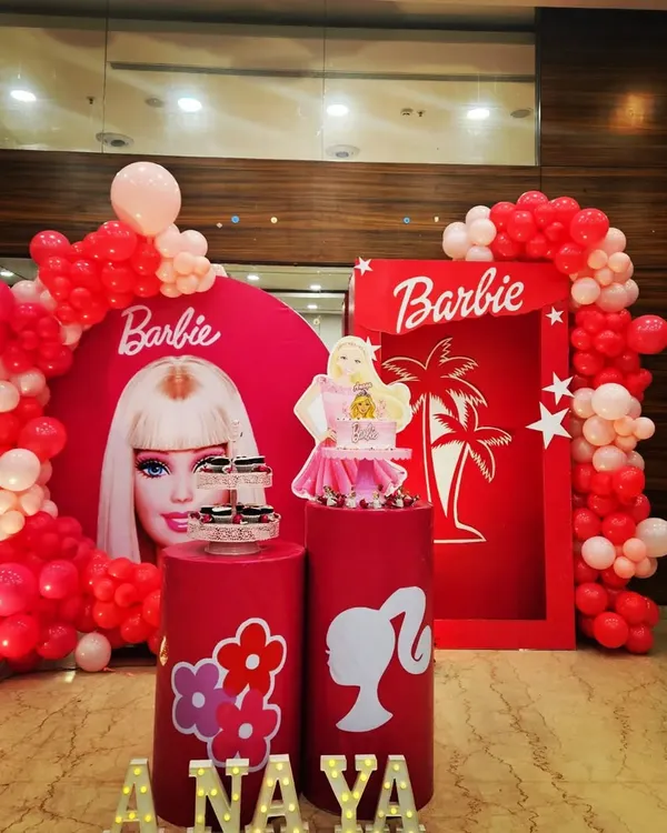 A classic Barbie theme for a fun birthday party. This setup features a round backdrop with the iconic Barbie face, a doll box photo prop, and a red and pink balloon garland.