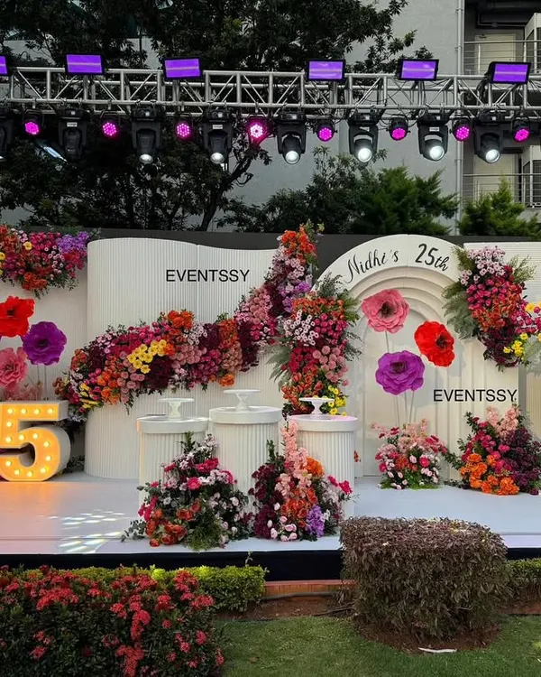 A wide shot of the stunning floral stage for a 25th birthday, featuring a clean white backdrop overflowing with colorful flowers and a large marquee number.