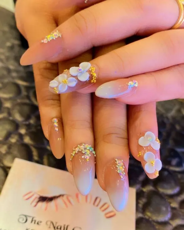 An intricate design featuring ombre nail extensions with sculpted 3D white flowers. Tiny crystals and glitter beads are added at the base and tips for a delicate, sparkling finish.