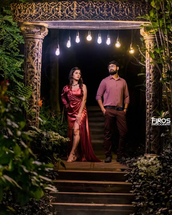 A moody and stylish night portrait. The couple stands under a row of Edison bulbs, creating a dramatic, low-light scene that feels both modern and intimate.